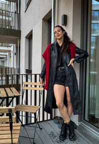 Μαύρο δερμάτινο μακρύ παλτό με κόκκινη πλεκτή λεπτομέρεια. Φοριέται πάνω από μαύρο πουκάμισο και σορτς, συνδυασμένο με μαύρες μπότες. Υπαίθρια μπαλκονάτη ρύθμιση.