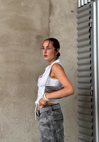 Bílý korzetový top s krajkovým zavazováním, šedé maskáčové cargo kalhoty, hřebíkovým černým páskem, průhlednými brýlemi a malou šedou kabelkou. Hladký pozadí zdi.