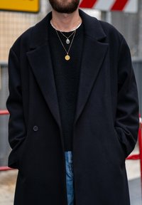 Cappotto di lana nera con ampi risvolti, indossato sopra un maglione nero, abbinato a jeans blu. Presenta collane d'oro a strati con pendenti.