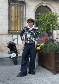 Black and white printed jacket with a collar, paired with wide-legged dark denim trousers and black shoes. Background features motorcycles and greenery.
