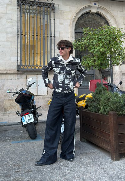 Chaqueta impresa en blanco y negro con cuello, combinada con pantalones de mezclilla oscuros de pierna ancha y zapatos negros. El fondo presenta motocicletas y vegetación.
