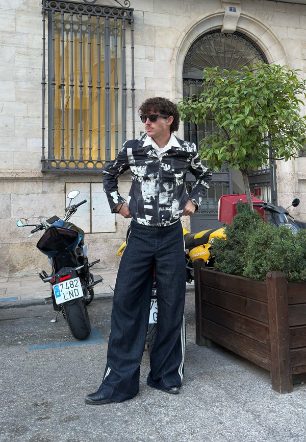 Black and white printed jacket with a collar, paired with wide-legged dark denim trousers and black shoes. Background features motorcycles and greenery.