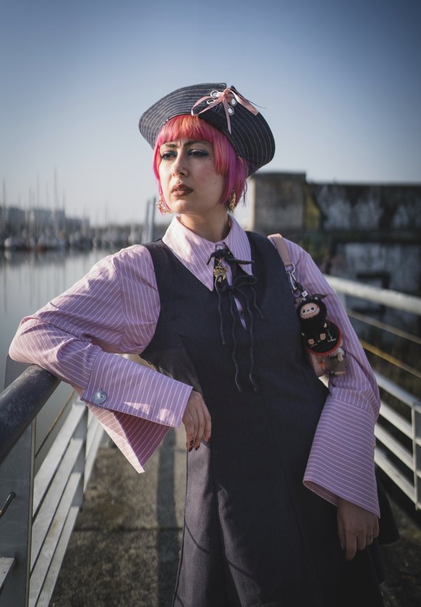 Person mit pinken Haaren und gestreiftem Hut lehnt am Geländer eines Yachthafens, trägt ein geschichtetes Outfit mit Weste, übergroßen Ärmeln und hält eine kleine Plüschtasche.