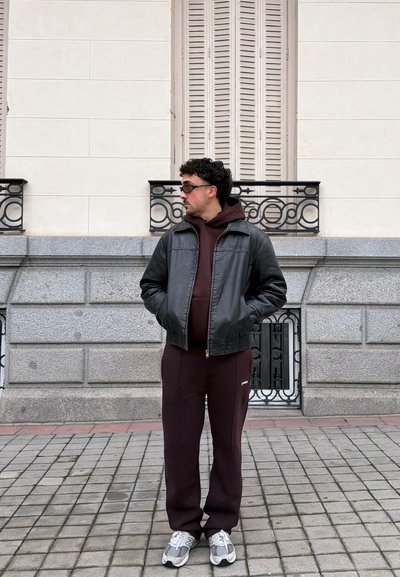 Hombre con chaqueta de cuero negra, sudadera con capucha burdeos y pantalones, zapatillas grises, de pie con las manos en los bolsillos frente a la fachada de un edificio de piedra.