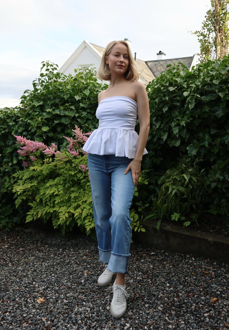 Blonde vrouw met een wit strapless topje en blauwe opgerolde spijkerbroek staat op een grindpad met groene struiken en bloemen achter haar.