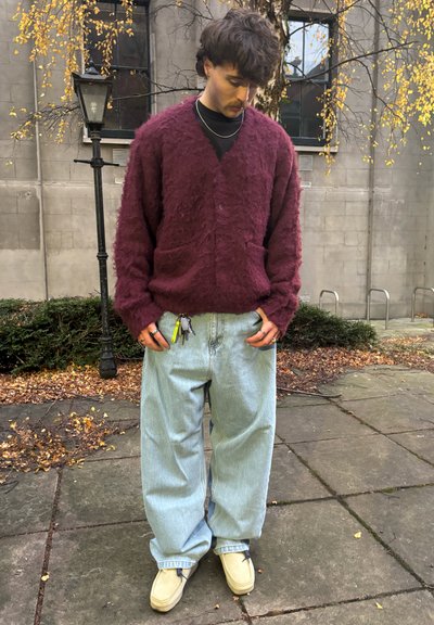 Cárdigan burdeos de felpa con escote en V, dos bolsillos laterales, combinado con jeans holgados de color azul claro y zapatos de tela color crema.
