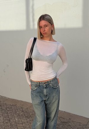 Joven con cabello corto y rubio que lleva una blusa blanca transparente de manga larga, pantalones vaqueros azules holgados y un bolso negro al hombro, frente a una pared lisa.