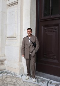 Rjav, dvoredni oversized blezer z ujemajočimi se hlačami, v kombinaciji z lahko puloverjem in belimi tenisicami. Majhna črna torbica na rami.