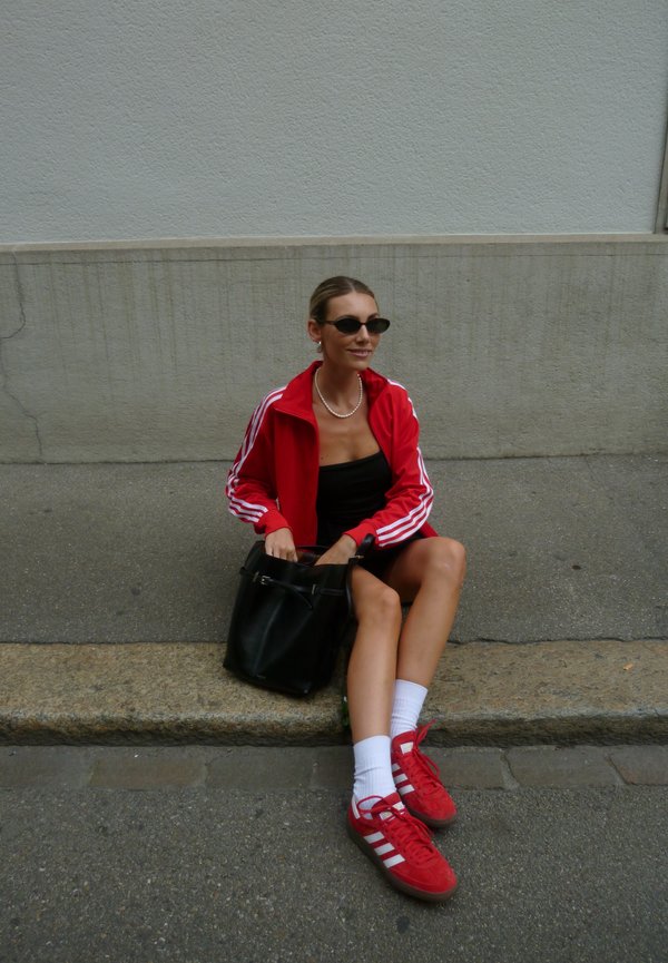 Trening roșu Adidas cu dungi albe, bluză neagră, geantă neagră, adidași roșii și șosete albe. Așezat pe un treaptă de piatră, sprijinit de un perete texturat.