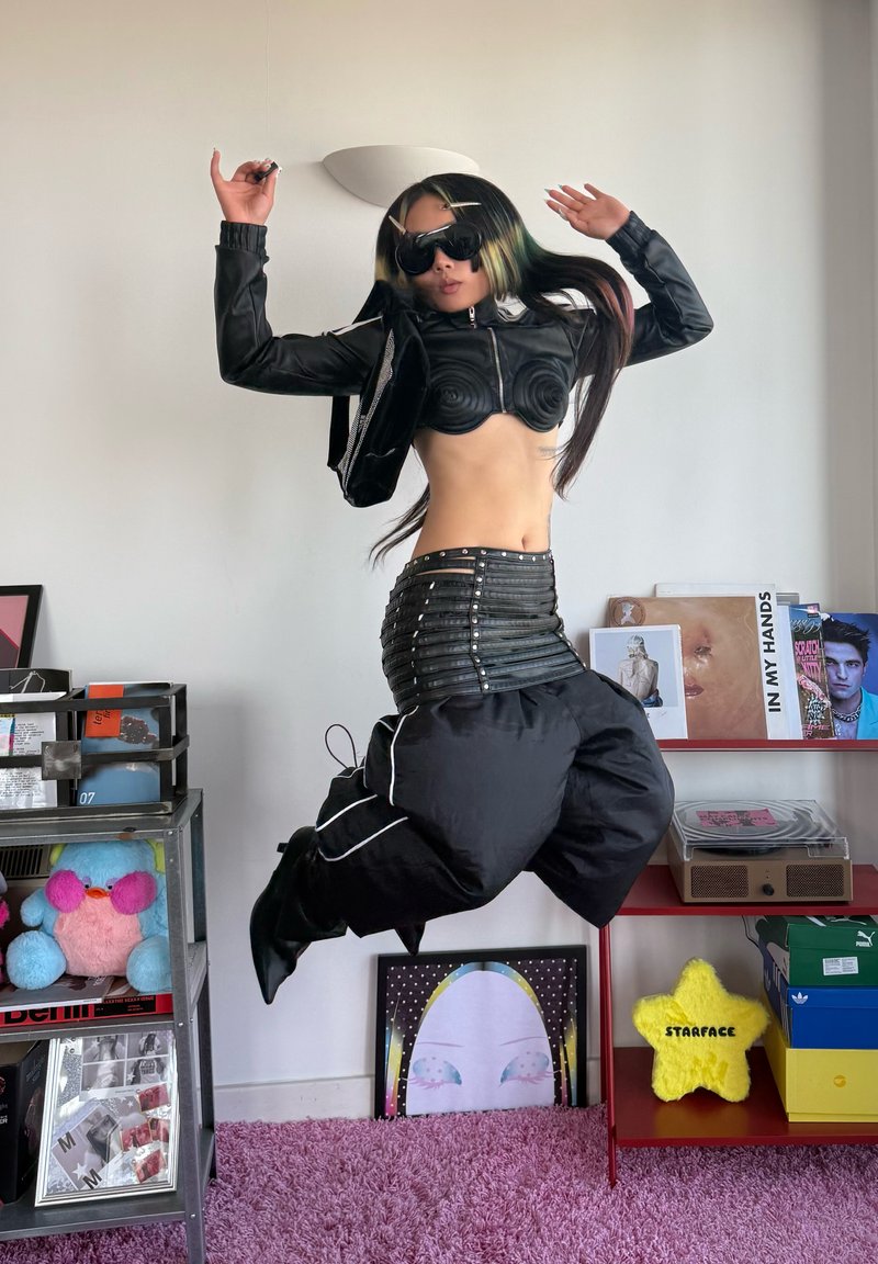 Personne portant un crop top en cuir noir, un pantalon noir large, des lunettes de soleil et des pinces saute dans une pièce avec des étagères, des peluches et des œuvres d'art encadrées.