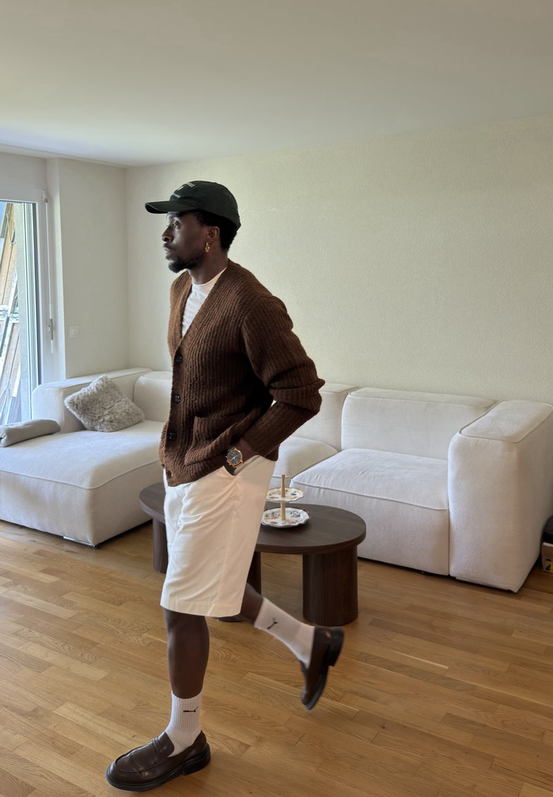 Homme portant un cardigan marron, un short blanc et des chaussures sombres marchant dans un salon moderne avec un canapé blanc et une table ronde en bois.