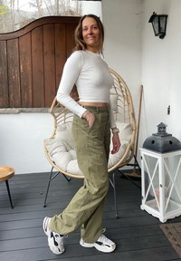 Χακί cargo παντελόνι με πλαϊνές τσέπες, άσπρη μακρυμάνικη cropped μπλούζα και χοντρά άσπρα sneakers. Σε φόντο με ανοιχτόχρωμο τοίχο και ξύλινη επιφάνεια.