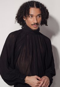 Haut plissé noir avec un col montant et des manches longues. Texture transparente avec un éclat subtil. Avec des poignets boutonnés et des pinces dans les cheveux.