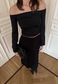 Femme portant un haut noir décolleté d'une épaule et une jupe longue assortie, tenant une pochette noire, debout sur un parquet en bois.