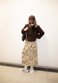 Femme portant une casquette marron, des lunettes de soleil foncées, un pull marron, une jupe camouflage beige, des baskets blanches et un sac à bandoulière noir à l'intérieur.