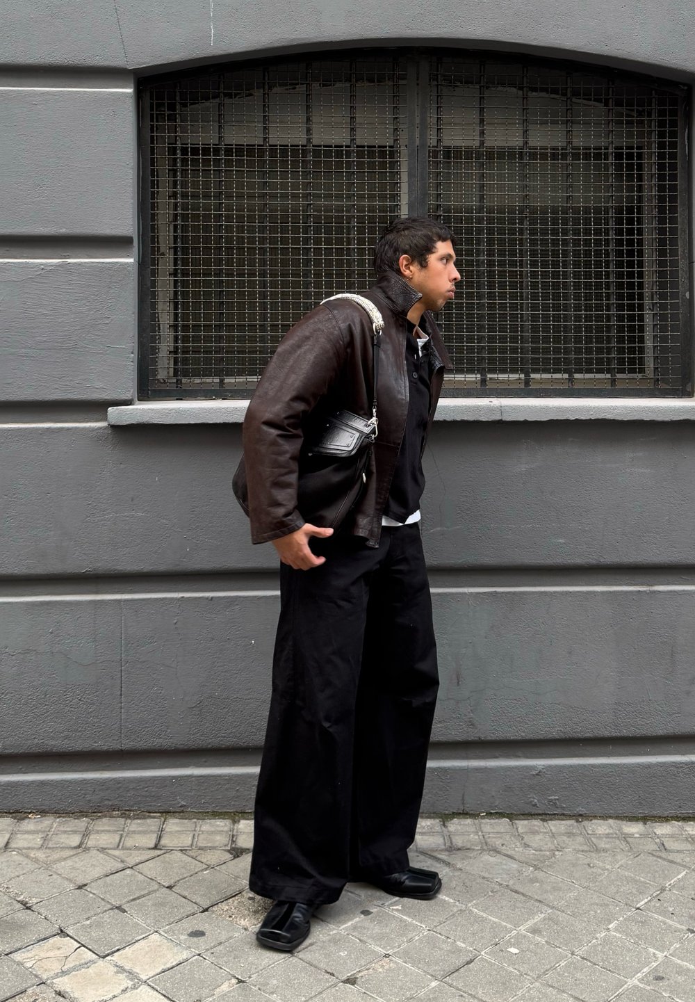 Καφέ δερμάτινο μπουφάν, μαύρο παντελόνι, μαύρα παπούτσια. Μικρή μαύρη τσάντα με ασημένιο λουρί. Γκρίζος τοίχος και παράθυρο με πλέγμα στο φόντο.