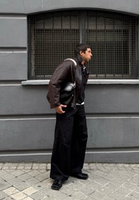 Καφέ δερμάτινο μπουφάν, μαύρο παντελόνι, μαύρα παπούτσια. Μικρή μαύρη τσάντα με ασημένιο λουρί. Γκρίζος τοίχος και παράθυρο με πλέγμα στο φόντο.