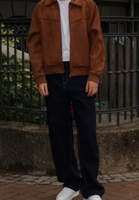 Giacca zip-up in suede sintetico marrone con colletto, abbinata a pantaloni larghi in denim scuro e sneakers bianche. Design pulito con cuciture a contrasto.