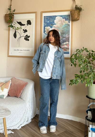 Chaqueta de mezclilla azul claro sobre una camiseta blanca, combinada con jeans acampanados azules. Zapatillas blancas. El fondo presenta plantas y obras de arte enmarcadas.