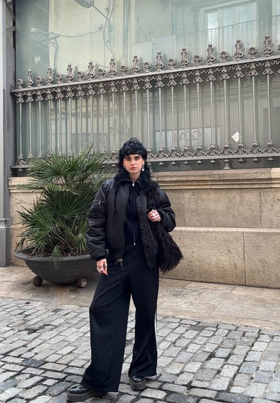 Chaqueta de plumón negra con cuello acanalado, suéter negro de punto, pantalones anchos negros y zapatos negros gruesos; sosteniendo un bolso negro texturizado.