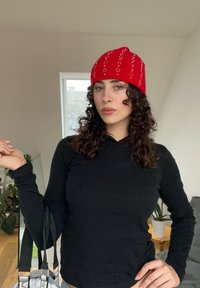 Bonnet en tricot rouge avec des rayures blanches verticales, associé à une chemise noire à manches longues. En arrière-plan, une plante et un mobilier moderne.