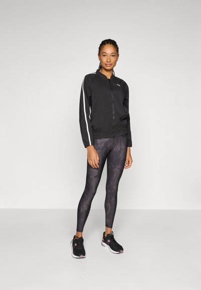 Chaqueta negra con cremallera y rayas blancas, combinada con leggings de patrones en negro y gris y zapatillas negras. Tejido texturizado, ajuste atlético.