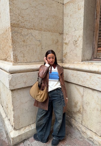 Joven mujer que lleva un abrigo de cuero marrón, suéter de rayas, vaqueros anchos, zapatos negros y bolso beige, de pie contra la esquina de una pared de piedra beige.