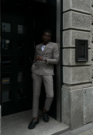 Traje de cuadros gris de doble botonadura con un corte ajustado, acompañado de mocasines negros. El fondo presenta una pared de piedra texturizada y una puerta oscura.