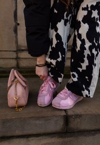 Baskets roses avec finition satinée et lacets en ruban. La tenue comprend un pantalon à motif vache noir et blanc. Un petit sac à main rose repose à proximité.