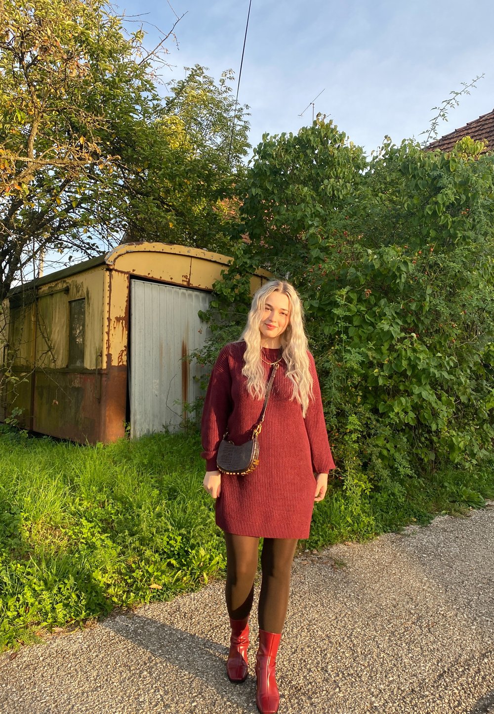 Strikket bordeaux kjole med lange ermer, svarte strømpebukser og blanke røde ankelstøvler. Liten crossbody-veske med teksturert design.