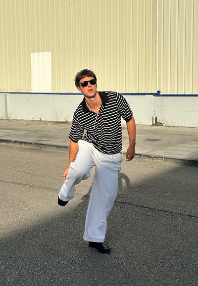 Camisa de polo a rayas blancas y negras, pantalones anchos blancos y zapatos negros. La persona está en una pose dinámica, usando gafas de sol oscuras.