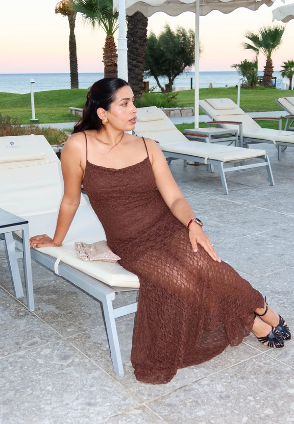 Una mujer con un vestido marrón de encaje está sentada en una tumbona junto al mar, con palmeras y tumbonas vacías detrás.