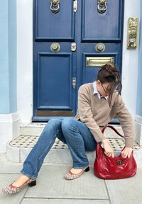 Červená kožená kabelka s kruhovým kovovým detailom, béžový sveter, modré džínsy, vzorované baleríny s blokovým podpätkom a modré dvere v pozadí.