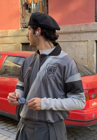 Tricou sportiv gri și negru cu logo în formă de scut, având dungi orizontale, guler și mâneci lungi cu dantelă. Ține ochelari de soare într-o mână.