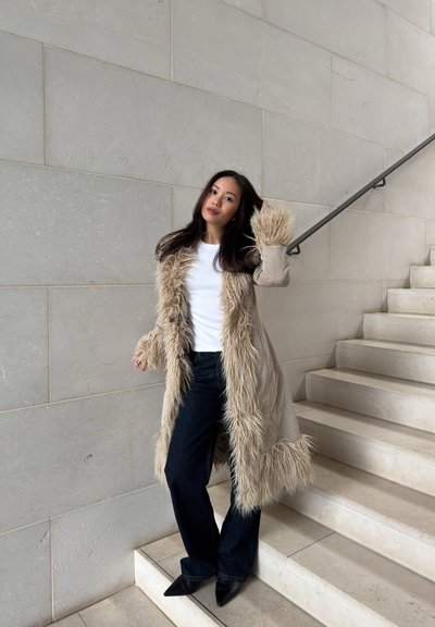 Mujer con abrigo beige con ribete de piel, camisa blanca y pantalones negros de pie en una escalera interior de piedra con una mano tocándose el pelo.