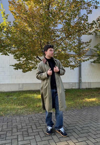 Abrigo largo verde oliva con cuello marrón, sudadera negra, jeans azules y zapatillas negras con cordones blancos, situado frente a un fondo de árboles.