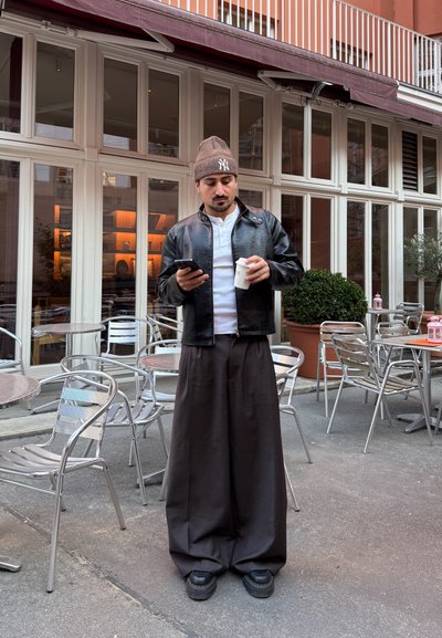 Chaqueta de cuero negra sobre una camisa blanca, pantalones anchos negros, gorro marrón, sosteniendo una taza de café, mirando un teléfono inteligente, en un café al aire libre.