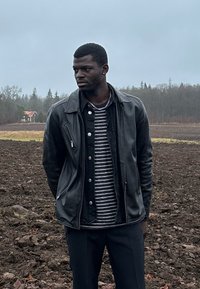 Homme portant une veste en cuir noire et une chemise rayée se tient dans un champ labouré, avec une petite maison rouge et des arbres en arrière-plan sous un ciel couvert.