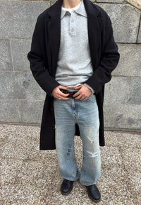 Άτομο που φοράει μαύρο παλτό, γκρι πλεκτό πόλο, φθαρμένο τζιν με φαρδιά μπατζάκια, μαύρα παπούτσια και ρυθμίζει τη ζώνη με ορατή μέση Calvin Klein.