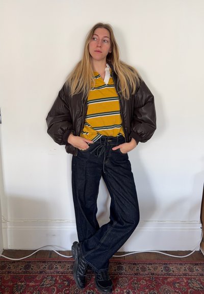 Chaqueta acolchada marrón, polo con rayas amarillas y verdes, vaqueros azul oscuro de pierna ancha y botines negros. Diseño sencillo, conjunto informal.