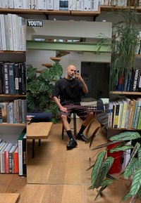 Spiegel die een persoon in een zwart shirt en bordeauxrode shorts reflecteert, zittend op een zwarte kruk, omringd door boekenplanken en planten.