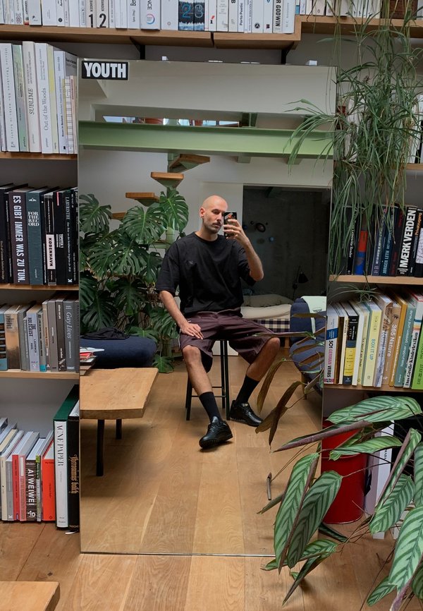 Spiegel die een persoon in een zwart shirt en bordeauxrode shorts reflecteert, zittend op een zwarte kruk, omringd door boekenplanken en planten.