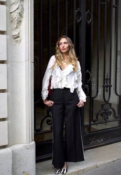 Mujer con cabello largo y ondulado, con blusa blanca con volantes, pantalones negros de pierna ancha y zapatos con estampado de cebra, está junto a una ornamentada puerta de metal negro.