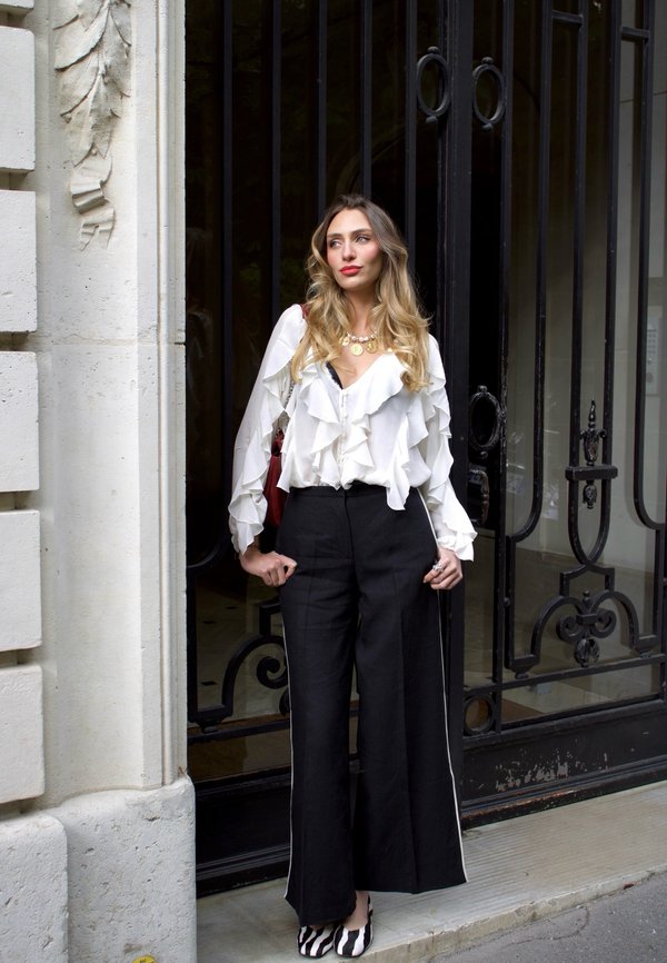 Vrouw met lang golvend haar, in een witte blouse met ruches, zwarte wijde broekspijpen en schoenen met zebraprint, staat bij een sierlijk zwart metalen hek.