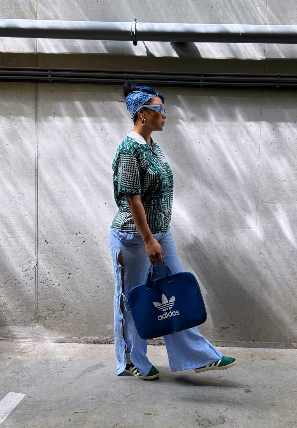 Camisa verde con estampado, pantalones acampanados de color azul claro con aberturas laterales, zapatillas deportivas verdes y blancas, y una mochila azul de Adidas. Gafas de sol grandes y diadema.