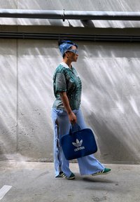 Camisa verde con estampado, pantalones acampanados de color azul claro con aberturas laterales, zapatillas deportivas verdes y blancas, y una mochila azul de Adidas. Gafas de sol grandes y diadema.