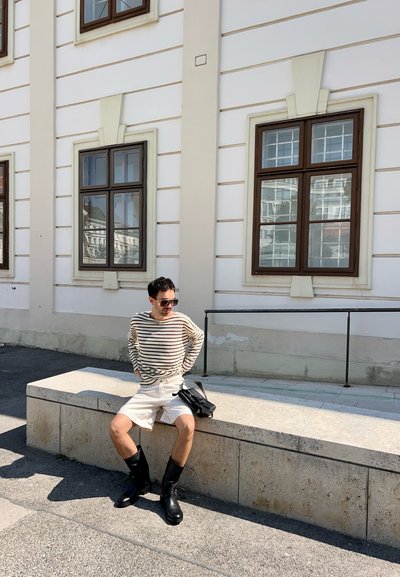 Camiseta de manga larga a rayas, pantalones cortos blancos, botas negras hasta la rodilla, sentada en un banco de piedra cerca de un edificio blanco con ventanas de madera.