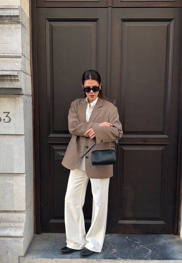 Mujer con blazer marrón oversize, pantalones blancos y zapatos negros, de pie con bolso negro frente a una puerta de madera oscura.