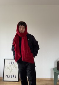 Rdeča šal s robovi, ovit okrog vratu, nošen z črno jakno in hlačami. V ozadju je cvetlični plakat in zelena mizica.