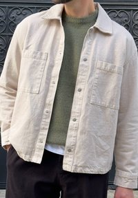 Man wearing a beige button-up jacket over a green knit sweater and black trousers, standing in front of an ornate dark metal door.